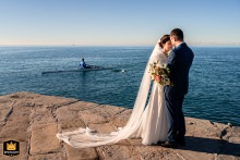 In Trieste, Italy, a canoe passes a stone jetty where a couple stands head-to-head during their wedding portraits.
