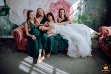 Bride in white wedding dress playfully lies across laughing, barefoot bridesmaids’ laps on pink velvet couch in floral-mural reception room.