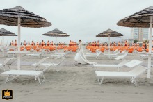 At Papeete Beach in Milano Marittima, Italy, the bride elegantly walks toward the sea for her beach wedding, gracefully passing between rows of empty lounge chairs that frame her serene journey.