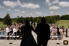 Silhouetted Bride and Escort Make a Dramatic Ceremony Entrance as Sunlit Guests Stand in Toronto The bride makes her ceremony entrance in Toronto, silhouetted with her escort in shadow. Guests stand by their seats, bathed in sunlight in the background.