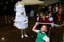Little Joy, Big Swing: An English Woodland Piñata Moment A young child joyfully swings a stick at a colorful, cake-shaped piñata. She is participating in a woodland wedding celebration in England.