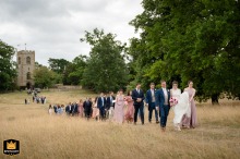 Bride and groom lead guests across a field at Courteenhall Estate, with the historic church visible in the background.