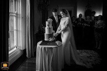 Black and white photo of a bride and groom cutting their wedding cake with window light at Kelmarsh Hall.