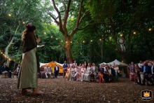 Heartfelt Vows and Golden Light Under the Woodland Canopy A wedding photographer captures heartfelt speeches under a canopy of trees at Woodland Weddings. The enchanting image shows guests listening intently, illuminated by warm strings of lights.