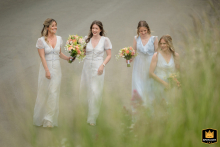 At St Peter & St Paul Church, Wing, Rutland, bridesmaids walk together along a dirt path bordered by tall green grass blades, the natural setting lending a fresh, serene backdrop to their procession.