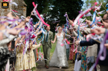 At All Saints Church, Yelvertoft, Northamptonshire, the bride and groom walk joyfully down the sunlit church path. Guests wave colorful ribbon wands, with sunlight and a shallow depth of field highlighting the grassy setting.