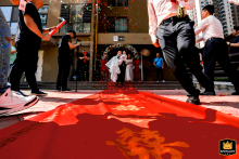From a low camera angle, the bride and groom walk down a red carpet as they leave the wedding venue in Taiyuan, Shanxi. Men nearby hurry about, adding dynamic energy to the celebratory scene.