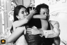 A black-and-white image at Four Seasons Bangkok shows a couple warmly hugging a woman, all three sharing an emotional embrace that expresses affection and heartfelt connection during the celebration.