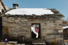 In a wintery scene in Alagna Valsesia, Italy, a bridesmaid is exiting a small stone building holding a dress