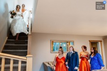 Bride descends rustic stairs at Hobbit Hill, Lancashire, warmly welcomed by smiling bridesmaids moments before her wedding ceremony.