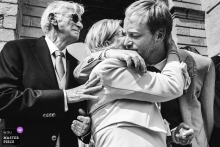 At Eglise d'Urrugne in Pyrénées Atlantiques, France, the photographer captures a moving low-angle black-and-white shot as the groom embraces his parents outside the church, expertly highlighting deep emotion and familial connection.