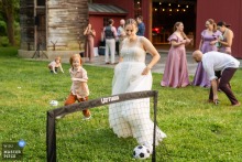 At The Intervale in Burlington, Vermont, the photographer captures the bride playing soccer with guests on the grass, red barn and silo in view, expertly highlighting celebration and lively outdoor interaction.