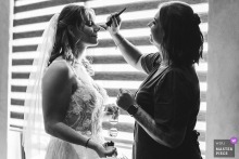 At Villa Dell'Angelo in Hildesheim, the bride receives a final touch-up before the ceremony, captured in black and white by a window with horizontal louvers behind her.