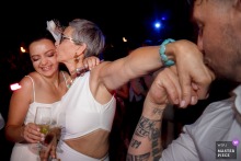 In Largentière, France, a woman offers her hand for a man to kiss while simultaneously kissing another woman’s cheek, sharing a playful double kiss during the wedding celebration.
