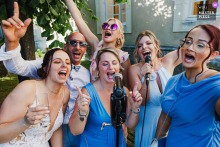 At a wedding reception in Moselle, a lively group of six guests—men and women, mostly in blue or white outfits—sing joyfully into a microphone, raising their arms and celebrating.
