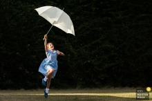 At a family farm reception venue in Northamptonshire, East Midlands, a flower girl joyfully plays with an umbrella against a dark background, in a minimalist frame full of childhood charm.