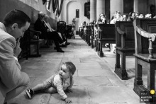 During the church ceremony, a young family member moves about while his father gently gestures for him to be quiet, capturing a candid and tender moment of parenting during the wedding.