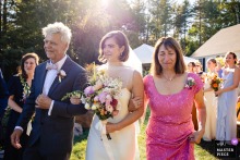   The bride’s parents escort her into the outdoor, sunny wedding ceremony in Houlton, Maine, radiating pride and happiness as she enters to begin this significant moment in her life.