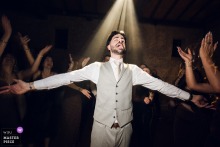 On the dance floor at the reception venue in Lyon, Rhône, France, the groom opens his arms to the sky as DJ light finds him, giving the moment a glow.