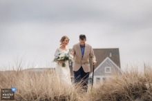 At the ceremony location in Gloucester Beach, Massachusetts, the bride walks with her father through coastal dune grass toward the groom, with a seaside home behind them and first sight nerves ahead.