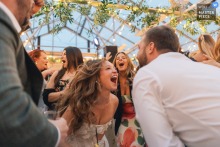 At Chapman's Greenhouse in Beverly, Massachusetts, the bride dances and sings with guests beneath the glass ceiling and hanging florals, the reception feeling bright, humid, and alive.