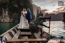 In Phuket, Thailand, the couple exchanges vows while the boat driver shifts the vessel back into the right background, a quietly comic reminder that even romance needs staging.