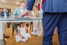 At City Hall in Dordogne, France, a little girl sits under the table bored through the civil ceremony, her blank expression providing a quietly humorous counterpoint to the formality around her.