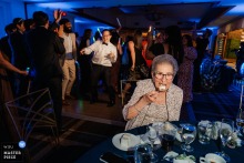 At Old Ranch Country Club in Seal Beach, California, the groom’s grandmother enjoys cake while the dance floor swirls beyond her, a sweet study in age, appetite, and timing.
