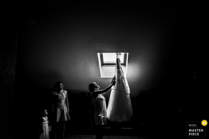 Une femme examine la robe de la mariée suspendue dans une lucarne sur cette photo en noir et blanc réalisée par un photographe de mariage du Piémont.