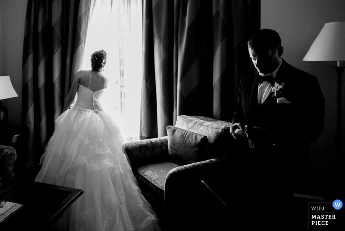 Schwarzweiss-Foto der Braut, die heraus ein Fenster als der Bräutigam schaut, überprüft seine Uhr von einem Hochzeitsfotografen San Antonio, TX.