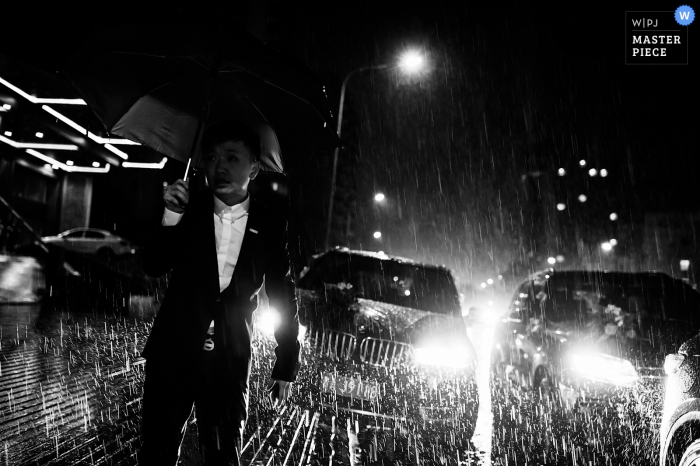 Photo: <a href="https://www.wpja.com/contests/2016-q3-photography-contest/weather/1913886"> Li Guo - Chine </a> Photo noir et blanc d'un homme debout sous la pluie, la nuit, avec deux véhicules derrière lui, par un photographe de mariage à Guizhou, en Chine.