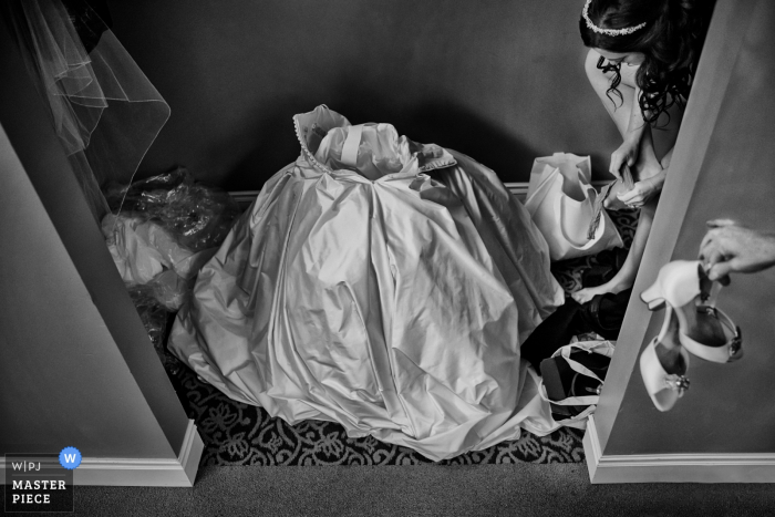 Photo by: <a href="https://www.wpja.com/contests/2016-q3-photography-contest/getting-ready/1895751">Ben Chrisman - United States</a> Black and white photo of the bride's dress on the floor ready for her to step into as a woman hands the bride her heels by a Charleston, SC wedding photographer.