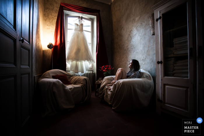 Photo by: <a href="https://www.wpja.com/contests/2016-q3-photography-contest/getting-ready/1910406">David Zaoui - United States</a> Photo of the bride sitting in a chair and looking up at her wedding dress hanging in the window by a Miami, FL wedding photographer.