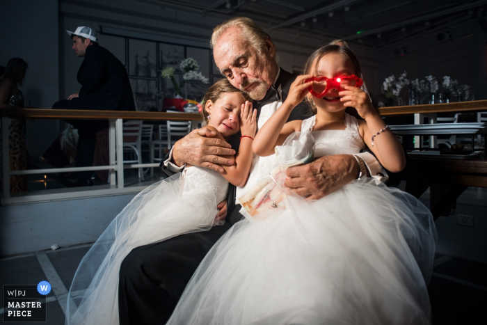 Foto van een man die twee jonge meisjes vasthoudt terwijl één speelt met een rode zonnebril, door een huwelijksfotograaf uit Madrid, Spanje.