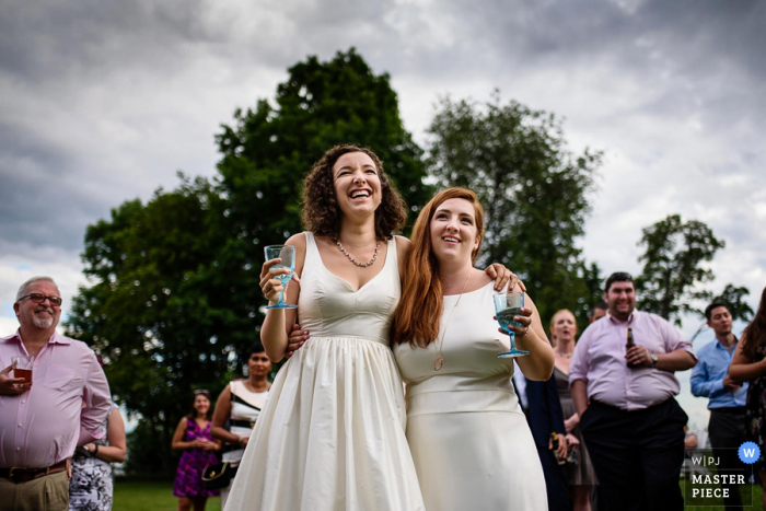 Two women stand with their arms around each other's shoulders as they hold their drinks Two women stand with their arms around each other's shoulders as they hold their drinks in this photo by a Burlington, VT wedding photographer.