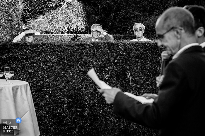 Photo by: <a href="https://www.wpja.com/contests/2016-q4-photography-competition/toastingspeeches/1963571">Philip Stephenson - France</a> Black and white photo of guests peering over a hedge to listen to the speech by a Paris wedding photographer.