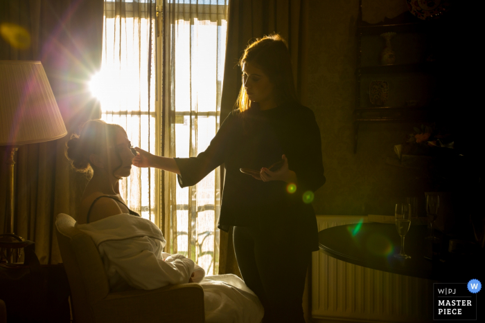 La sposa si siede a truccarsi mentre il sole splende attraverso la finestra in questa foto di un fotografo di reportage di matrimonio dell'Essex, in Inghilterra.