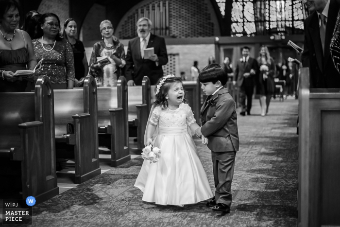 Foto di: <a href="https://www.wpja.com/contests/2016-q4-photography-competition/kids-being-kids/1982926"> Abram Eric Landes - Stati Uniti </a> Foto in bianco e nero di un giovane portatore dell'anello che aiuta la ragazza di fiore lungo il corridoio da un fotografo di matrimoni di Washington, DC.