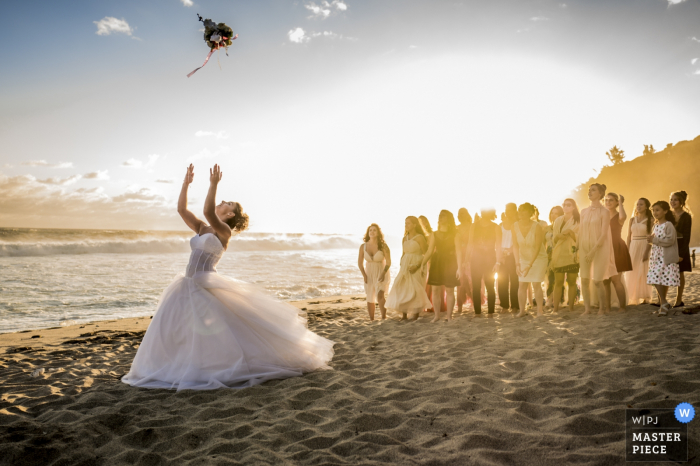Photo by: <a href="https://www.wpja.com/contests/2016-q4-photography-competition/bouquetgarter-action/1936676">Alexandre Kauder - Switzerland</a> The bride and guests stand on a sunny beach as she tosses her bouquet over her head in this photo by a Geneva, Switzerland wedding photographer.