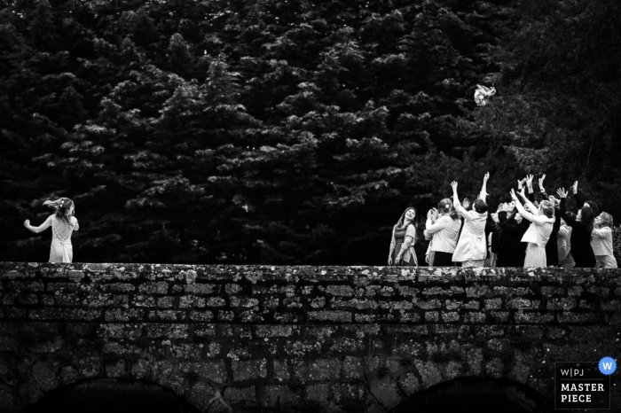 Schwarz-Weiß-Foto eines französischen Hochzeitsfotografen, auf dem die Braut ihren Blumenstrauß auf eine Steinbrücke wirft.