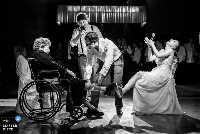 Photo by: <a href="https://www.wpja.com/contests/2016-q4-photography-competition/bouquetgarter-action/1965046">Andres Preumayr - Argentina</a> A young man puts the bride's garter on an older woman as the bride laughs in this black and white photo by a Santa Fe, Argentina wedding photographer.