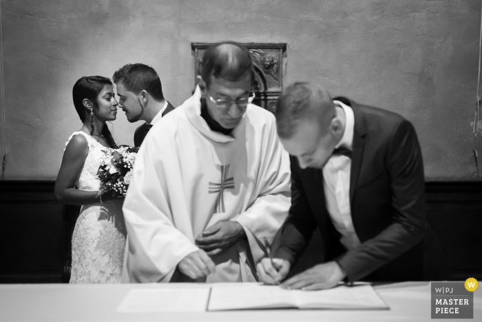 Photo par: <a href="https://www.wpja.com/contests/2016-q4-photography-competition/ceremony/1934346"> Louise Querin - France </a> Le témoin appose sa signature sur le prêtre tandis que la mariée et le marié se tiennent à l'arrière-plan sur cette photo en noir et blanc d'un photographe de mariage français.