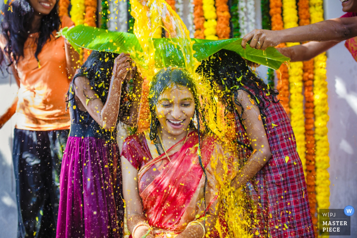 Foto von: <a href="https://www.wpja.com/contests/2016-q4-photography-competition/ceremony/1951516"> Alpheus Danson - Indien </a> Zwei Mädchen halten ein großes Schutzblatt hoch, während die Braut auf diesem Foto von einem indischen Hochzeitsfotografen mit einer gelben Flüssigkeit übergossen wird.