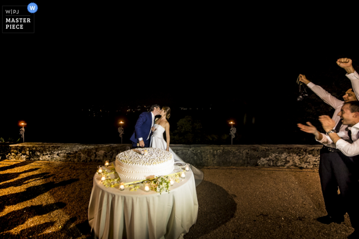 Foto door: <a href="https://www.wpja.com/contests/2016-q4-photography-competition/cake-cutting/1963901"> Luigi Rota - Italië </a> De bruid en bruidegom kussen voor hun bruidstaart terwijl hun gasten juichen op deze foto door een huwelijksfotograaf uit Lombardije, Italië.