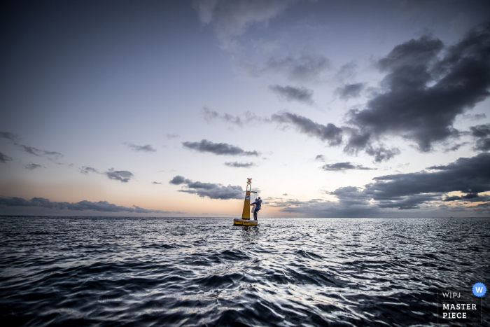 This distance shot of a bride and groom standing on a buoy in the ocean at sun set was captured by a Miami wedding photographer