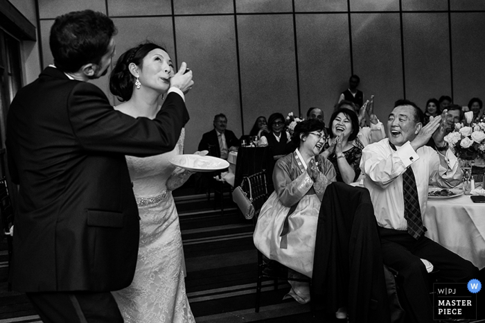 Il fotografo di matrimoni di Carson City ha catturato questa immagine giocosa in bianco e nero di uno sposo che allatta la torta della sposa con una forchetta mentre gli invitati al matrimonio li incoraggiano