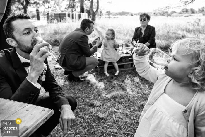 El fotógrafo de bodas de Montpellier creó esta imagen en blanco y negro de una niña y un padrino de boda que comparten un brindis mientras se sientan en mesas de picnic en un campo de hierba.