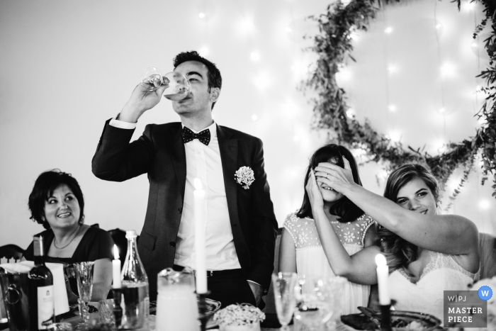 Il fotografo di matrimoni di Porto ha creato questa immagine in bianco e nero della sposa e dello sposo alla reception, mentre lo sposo brinda e la sposa copre scherzosamente gli occhi delle ragazze di fiore per proteggerla da qualcosa fuori dalla fotocamera