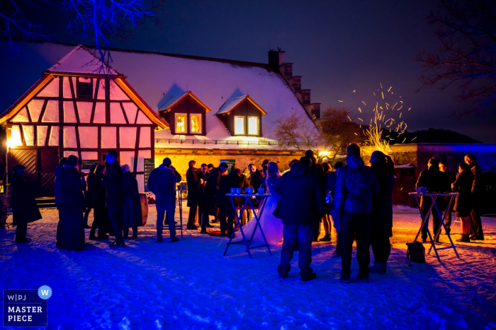 Der Hochzeitsfotograf aus Offenburg hat dieses Foto einer schneebedeckten Landschaft aufgenommen, während sich die Gäste außerhalb einer Villa schlängeln und sich um ein Feuer scharen