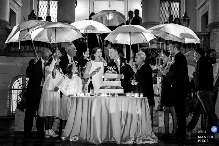 Der toskanische Hochzeitsfotograf hat dieses Schwarzweißfoto der Braut und des Bräutigams aufgenommen, die ihre Hochzeitstorte unter einem improvisierten Dachschutz schneiden